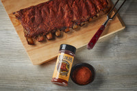 Rack of barbecued ribs on a wooden carving board with a Wildlife Seasonings rub bottle and a bowl of red rub