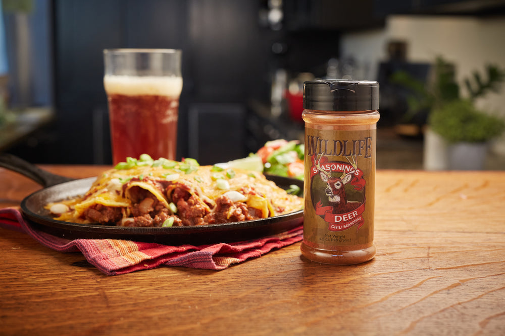 Skillet of venison enchiladas with a glass of beer and a Wildlife Seasonings Deer Chili Seasoning bottle on a wood table